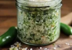 Homemade jalapeño salt in a glass jar with fresh jalapeños on the side.